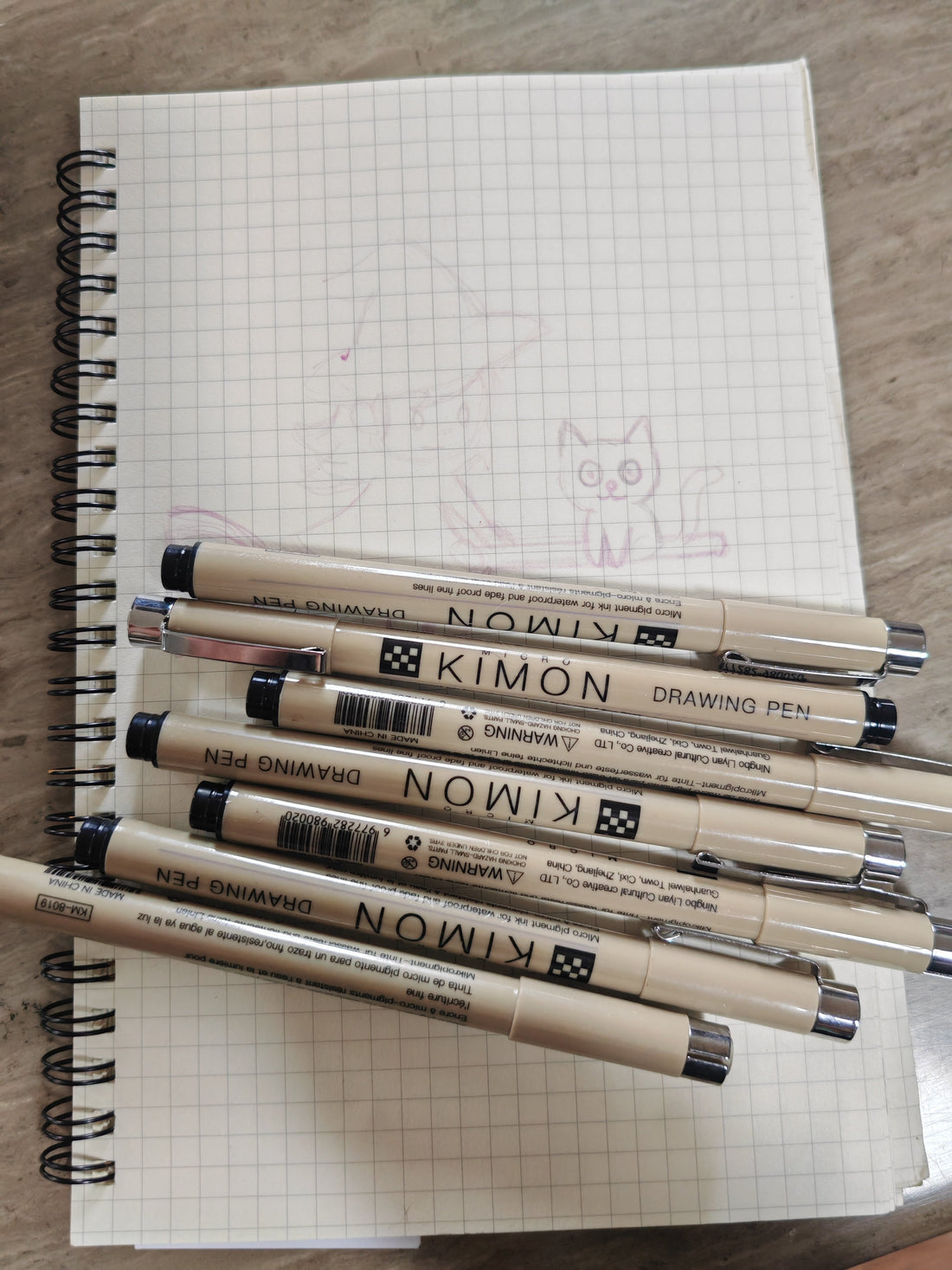 A photograph of a notepad featuring a sketch of a witch and a cat riding a broomstick.  A selection of black fine liners sit on top of the pad.