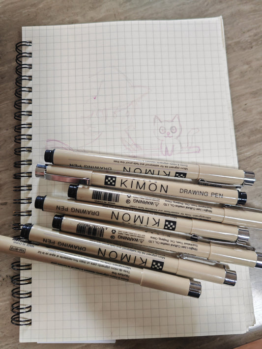 A photograph of a notepad featuring a sketch of a witch and a cat riding a broomstick.  A selection of black fine liners sit on top of the pad.